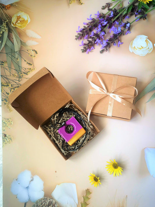 Handmade Purple and Golden Ceramic Square Necklace Pendant On a Bleck String Inside a Gift Box Surrounded By Wildflowers